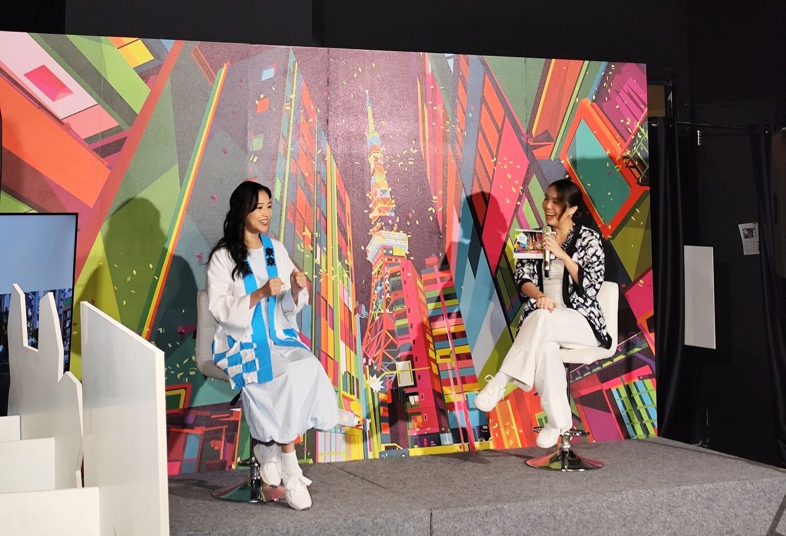 Talkshow press conference Walk Tokyo, bintang tamu berbaju happi biru putih dan MC duduk di kursi tinggi di depan backdrop ilustrasi Tokyo Tower