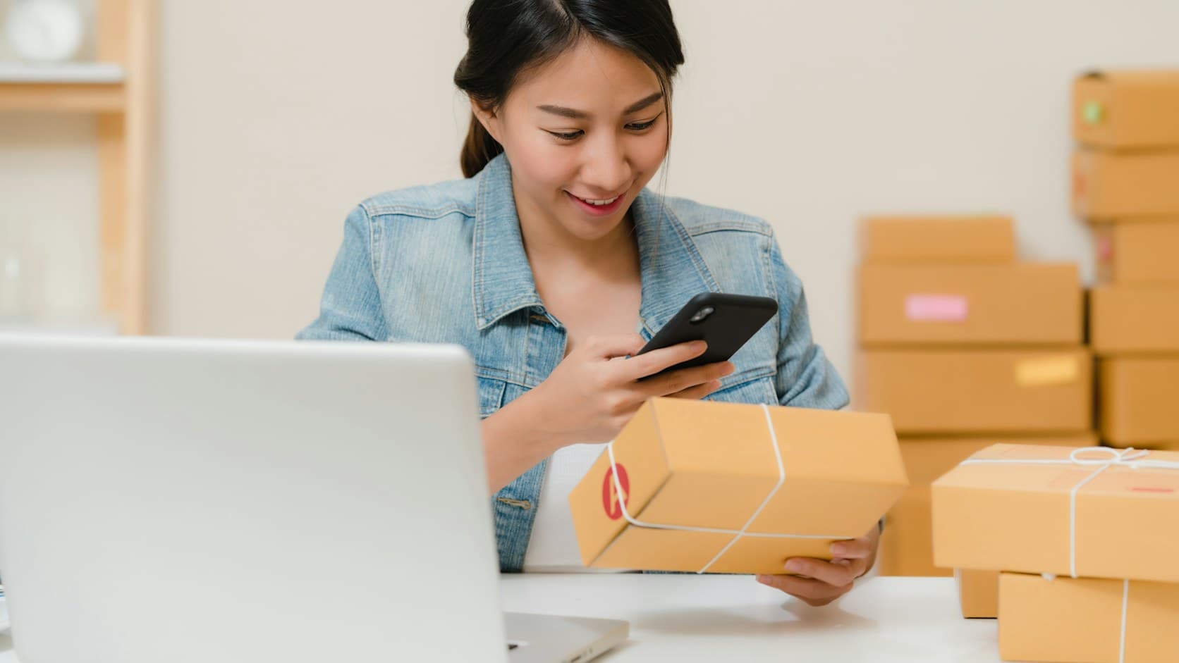 Pemilik toko online tersenyum memotret paket dengan ponsel di dekat laptop dan tumpukan kardus