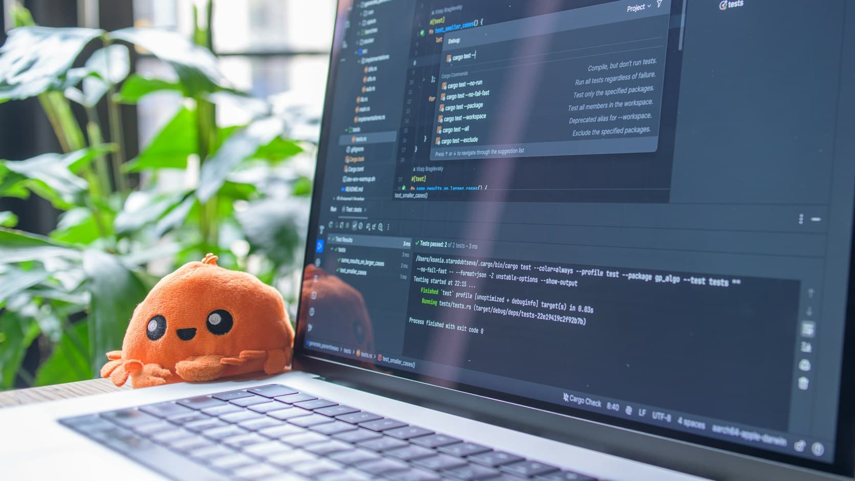 Laptop yang menampilkan editor kode dan terminal, dengan boneka kepiting oranye kecil di sampingnya dan tanaman di belakang