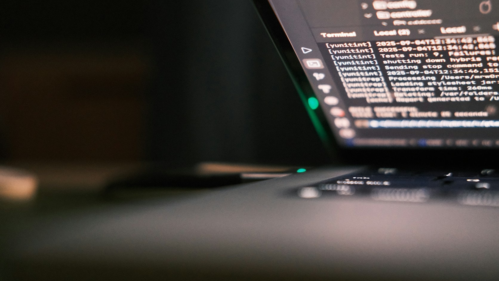 Layar laptop gelap menampilkan baris-baris log di jendela terminal, difoto dari samping