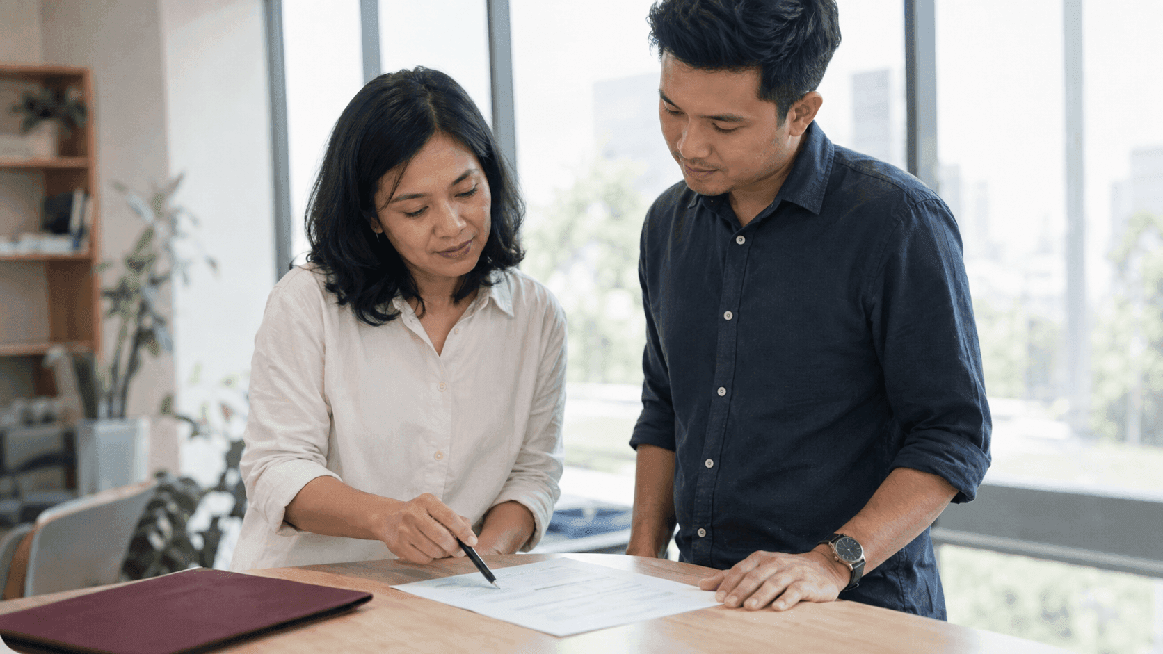 Dua rekan kerja meninjau laporan cetak bersama di meja kantor yang terang, perempuan memegang pulpen dan menunjuk satu baris sementara laki-laki melihat dengan saksama, jendela besar di belakang mereka