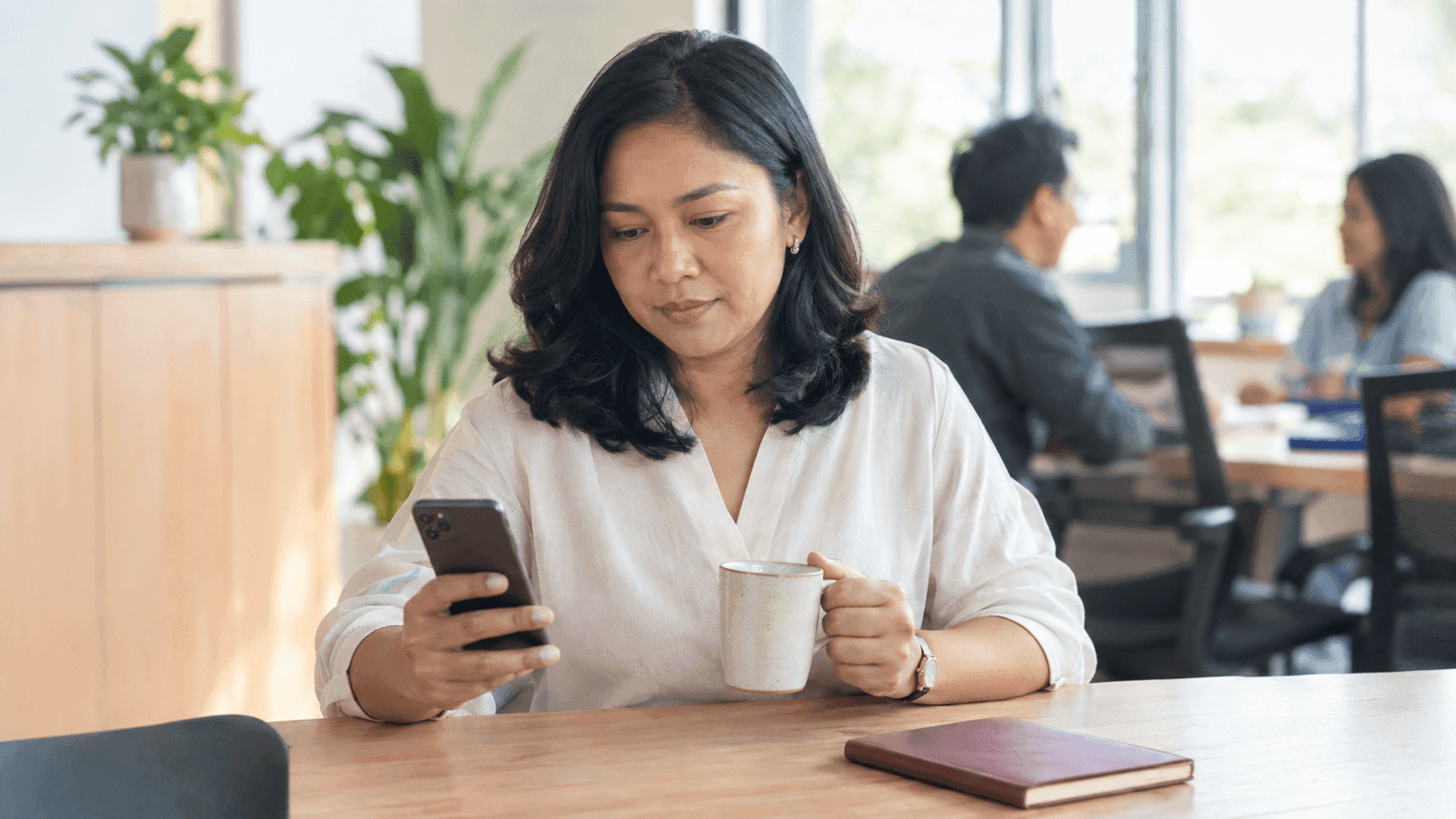 Pemilik usaha perempuan duduk di pantry kantor yang terang di pagi hari, membaca ponsel sambil memegang cangkir kopi, dengan dua rekan kerja berbincang di latar belakang yang buram