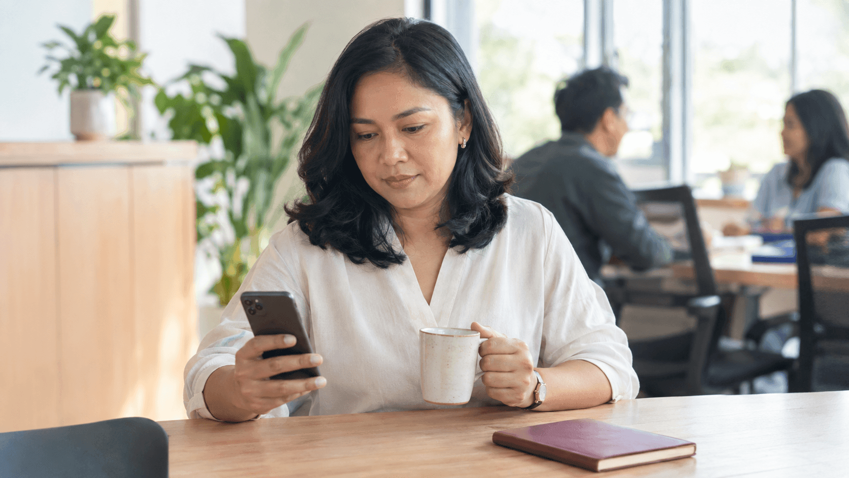 Pemilik usaha perempuan duduk di pantry kantor yang terang di pagi hari, membaca ponsel sambil memegang cangkir kopi, dengan dua rekan kerja berbincang di latar belakang yang buram