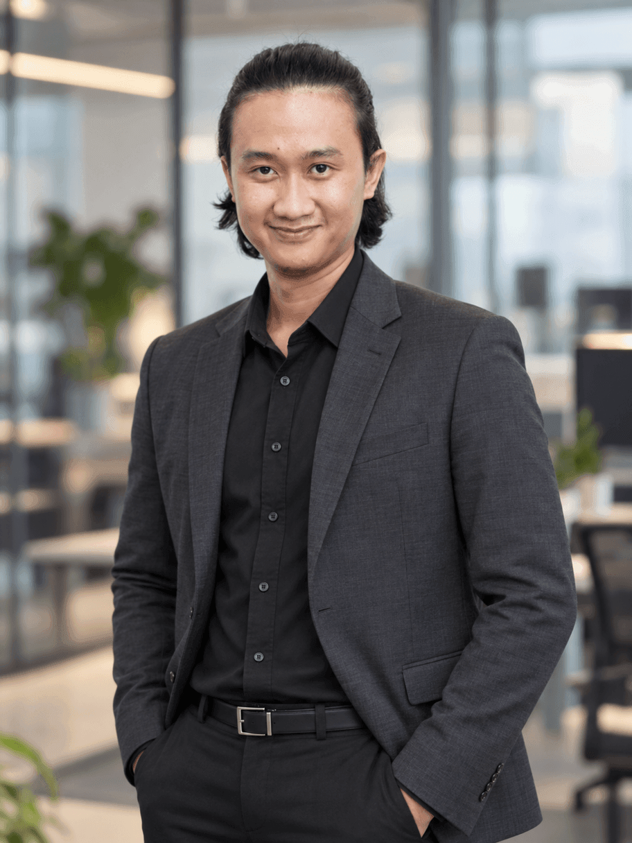 Hasil AI gaya konsultan: pria yang sama memakai blazer charcoal dan kemeja hitam, tangan di saku, berdiri di kantor modern berdinding kaca yang blur