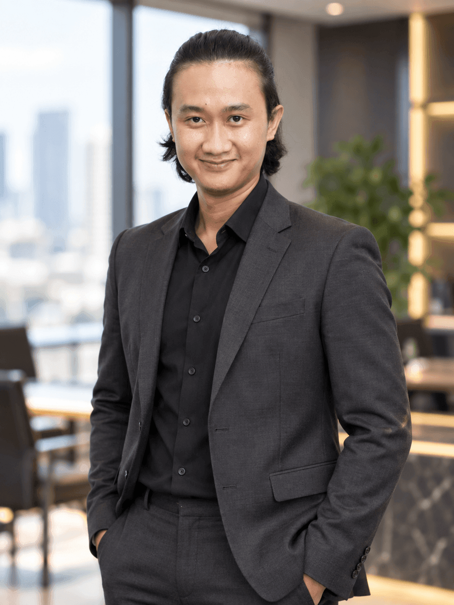 Hasil AI gaya eksekutif: pria yang sama memakai setelan charcoal dan kemeja hitam tanpa dasi, berdiri di ruang kantor kelas atas dengan jendela menghadap gedung kota yang blur