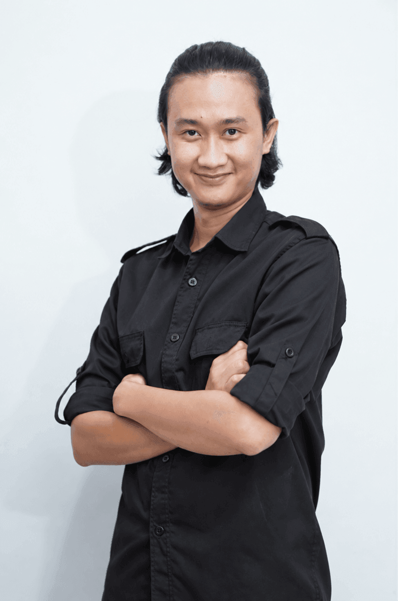 Foto studio seorang pria berambut panjang diikat, memakai kemeja hitam lengan digulung, tangan bersilang, latar putih polos dengan bayangan lembut